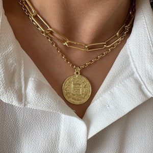 May include: A gold chain necklace with a large gold coin pendant featuring an intricate design. The necklace is layered over a second, thinner gold chain.