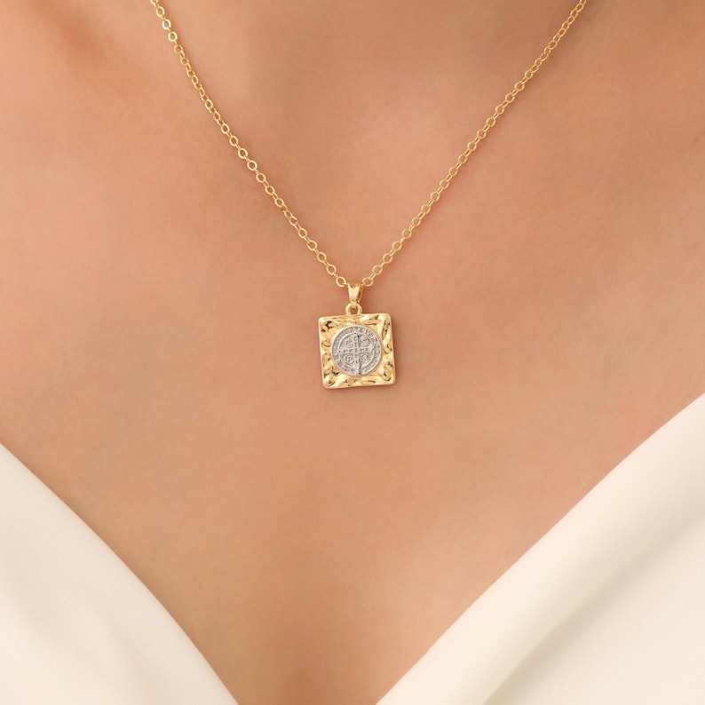 May include: A gold chain necklace with a square pendant featuring a circular design with a cross inside. The pendant has a textured, hammered finish.