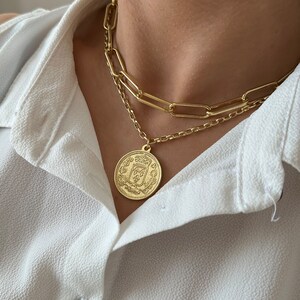 May include: A gold chain necklace with a large, round pendant featuring an intricate design. The pendant is a gold coin with a crest and other details.