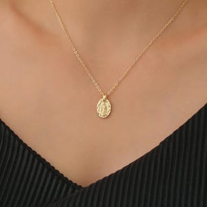 Pode incluir: Um colar de corrente de ouro com um pequeno pingente oval que apresenta uma imagem religiosa. O pingente tem uma superf&iacute;cie texturizada.