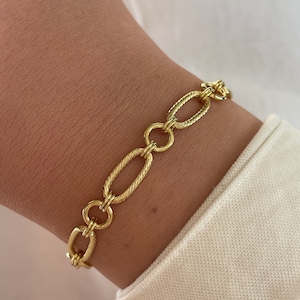 May include: A gold-colored chain bracelet with oval and circular links. The bracelet has a textured design on the oval links. The bracelet is worn on a wrist, with a white shirt sleeve in the background.
