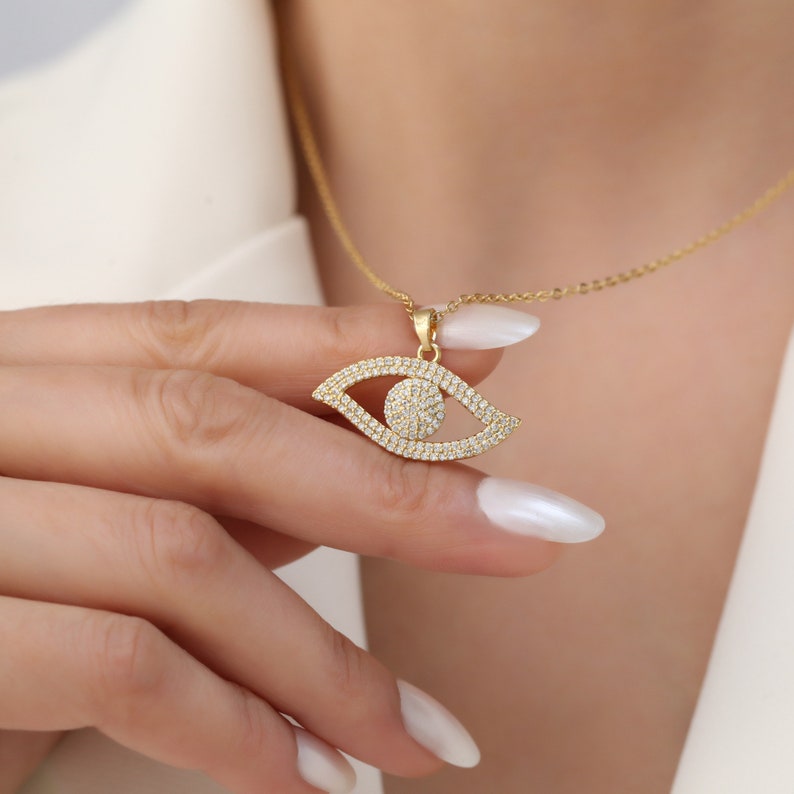 May include: Gold necklace with a large, eye-shaped pendant encrusted with clear cubic zirconia stones.