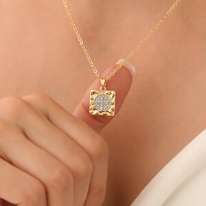 May include: Gold chain necklace with a square pendant featuring a round, textured, silver-toned medallion with a design in the center.
