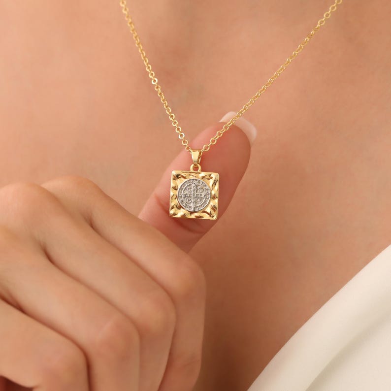 May include: Gold chain necklace with a square pendant featuring a round, textured, silver-toned medallion with a design in the center.