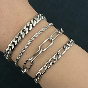 May include: Four silver bracelets of different designs are stacked on a wrist. The bracelets include a thick curb chain, a rope chain, a link chain, and a thinner curb chain. The bracelets are made of a shiny, metallic material.