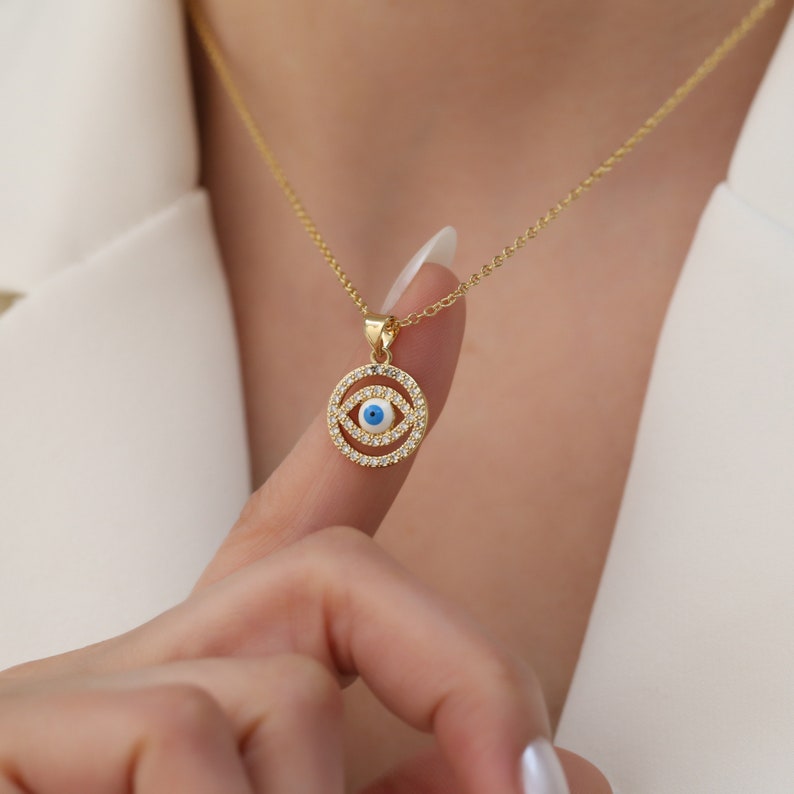 May include: A gold necklace with a round pendant featuring a blue evil eye with white and blue accents surrounded by clear sparkling stones.