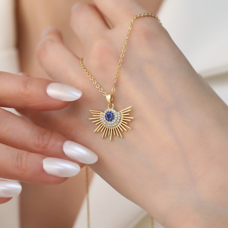May include: A gold necklace with a blue and white evil eye pendant. The pendant is surrounded by a sunburst design.