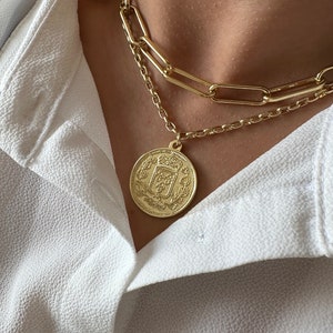 May include: A gold chain necklace with a large, round, gold coin pendant featuring an intricate design. The necklace is layered with a thicker, chunky chain.