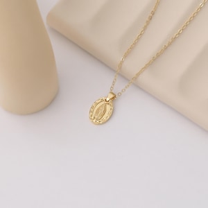 Pode incluir: Um colar de corrente dourada com um pequeno pingente oval que apresenta uma imagem detalhada da Virgem Maria.