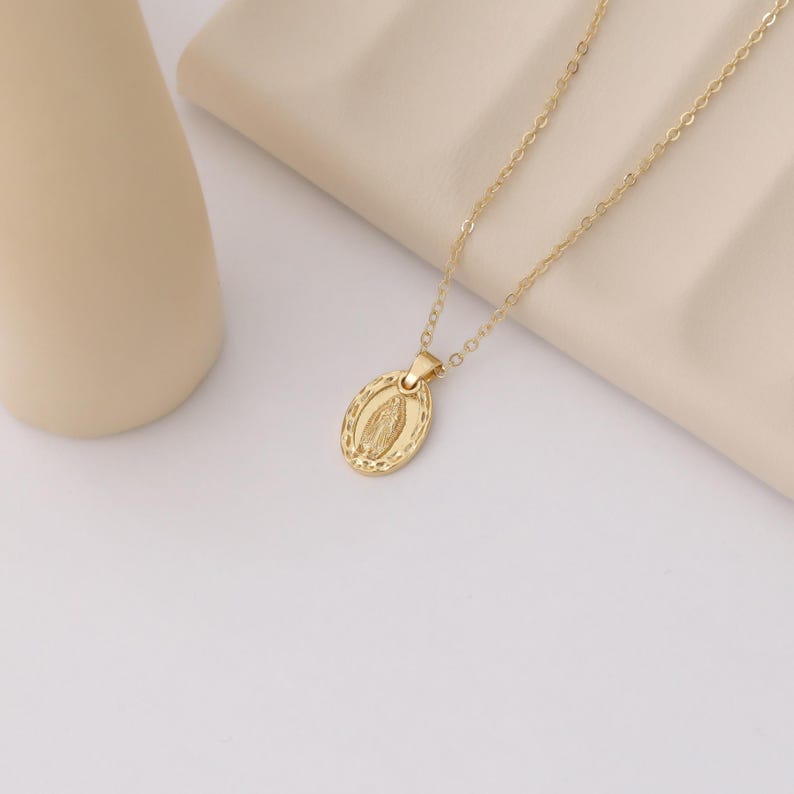 Pode incluir: Um colar de corrente dourada com um pequeno pingente oval que apresenta uma imagem detalhada da Virgem Maria.