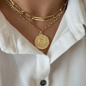 May include: A gold chain necklace with a large gold coin pendant featuring an intricate design. The necklace is layered over a second, thinner gold chain.