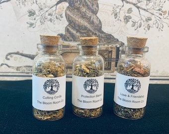 May include: Three small glass bottles with cork stoppers filled with dried herbs. Each bottle has a white label with a tree logo and text. The labels read "Cutting Cords The Bloom Room Co", "Protection Blend The Bloom Room Co", and "Love & Friendship The Bloom Room Co".