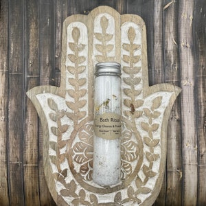 May include: A clear glass tube filled with white bath salts and dried herbs, with a silver lid. The tube is labeled "Bath Ritual Energy Cleanse & Protect". The tube rests on a carved wooden hand-shaped decorative piece.