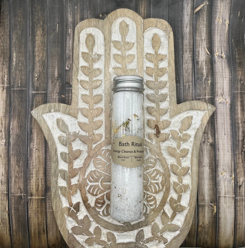 May include: A clear glass tube filled with white bath salts and dried herbs, with a silver lid. The tube is labeled "Bath Ritual Energy Cleanse & Protect". The tube rests on a carved wooden hand-shaped decorative piece.