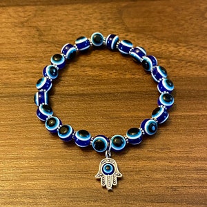 Puede incluir: Una pulsera de cuentas azules y blancas con diseño de ojo turco. Tiene un dije plateado en forma de mano, también con un diseño de ojo turco. La pulsera está sobre una superficie de madera.