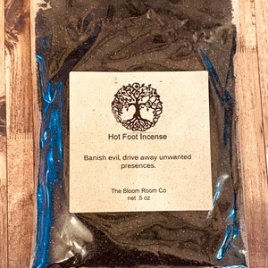 Op de afbeelding: Een klein plastic zakje gevuld met zwart poeder met het label "Hot Foot Incense". Het label vermeldt ook "Banish evil, drive away unwanted presences." en "The Bloom Room Co net .5 oz".