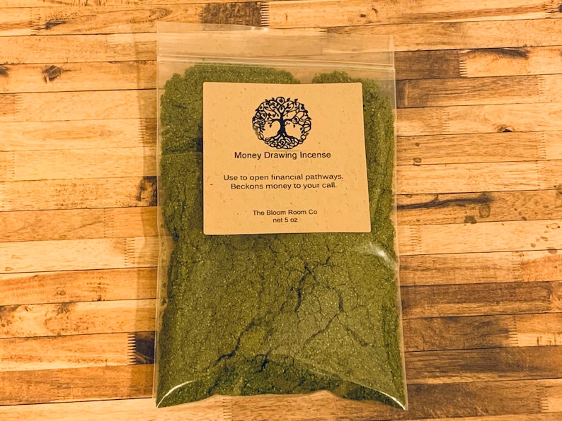 Op de afbeelding: Een kleine doorzichtige plastic zak met groene wierook met een bruin papieren etiket met de tekst "Money Drawing Incense" en de tekst "Use to open financial pathways, Beckons money to your call. The Bloom Room Co net 5 oz."