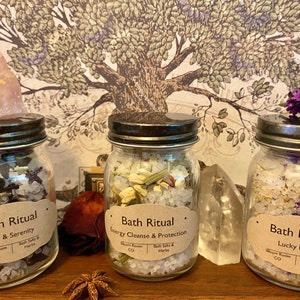 May include: Three glass jars filled with bath salts and herbs, labeled "Bath Ritual." The jars are topped with crystals and flowers. The labels read "Love & Serenity," "Energy Cleanse & Protection," and "Lucky Mojo."