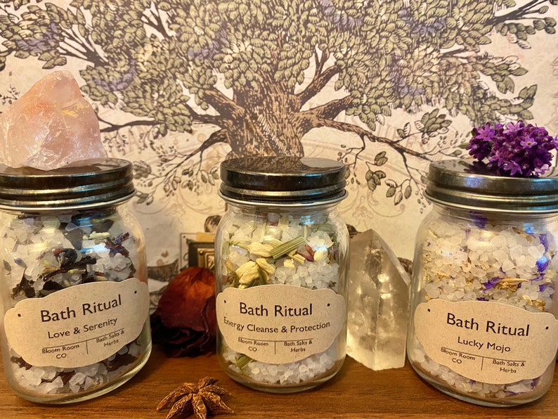 May include: Three glass jars filled with bath salts and herbs, labeled "Bath Ritual." The jars are topped with crystals and flowers. The labels read "Love & Serenity," "Energy Cleanse & Protection," and "Lucky Mojo."