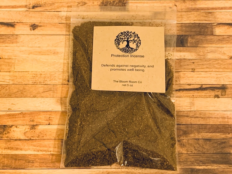 Op de afbeelding: Een bruine papieren zak gevuld met losse groene wierook. De zak heeft een label met de tekst "Protection Incense" en "Beschermt tegen negativiteit en bevordert het welzijn." Het label bevat ook de bedrijfsnaam "The Bloom Room Co" en het nettogewicht "5 oz".