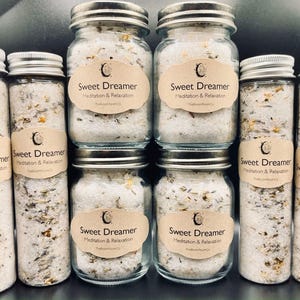 May include: An assortment of glass jars and tubes filled with bath salts. Each container has a silver lid and a label that reads "Sweet Dreamer Meditation & Relaxation." The salts are white with visible herbs and flowers.