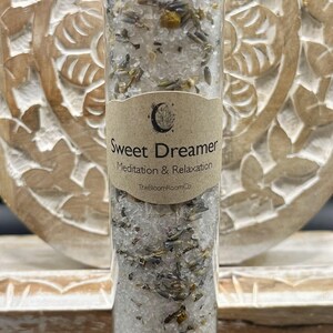 May include: A clear glass tube filled with white granules, dried herbs, and flowers, topped with a silver lid. A brown label reads "Sweet Dreamer Meditation & Relaxation". The product is from TheBloomRoomCo.