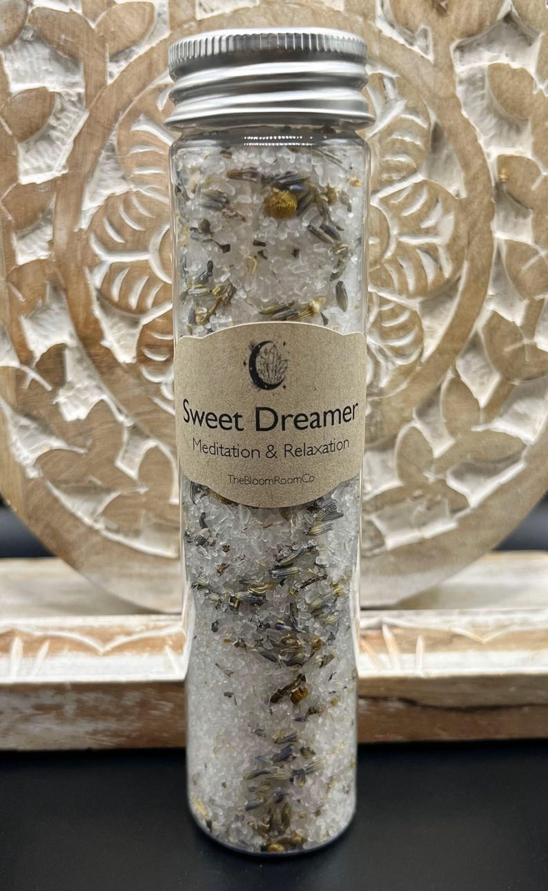 May include: A clear glass tube filled with white granules, dried herbs, and flowers, topped with a silver lid. A brown label reads "Sweet Dreamer Meditation & Relaxation". The product is from TheBloomRoomCo.