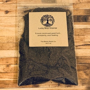 Op de afbeelding: Een bruine papieren zak met een label waarop staat "Lucky Mojo Incense" en "Ensure continued good luck, prosperity, and healing." Het label bevat ook de tekst "The Bloom Room Co" en "net 5 oz". De zak bevat zwarte wierook.
