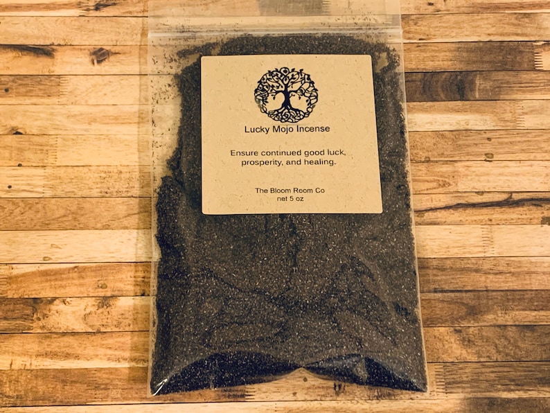 Op de afbeelding: Een bruine papieren zak met een label waarop staat "Lucky Mojo Incense" en "Ensure continued good luck, prosperity, and healing." Het label bevat ook de tekst "The Bloom Room Co" en "net 5 oz". De zak bevat zwarte wierook.