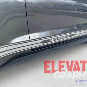 K&ouml;nnte beinhalten: Ein grauer Ford Mustang mit einem schwarzen und grauen Streifenaufkleber an der Seite. Der Aufkleber enth&auml;lt das Wort "Mustang" und ein stilisiertes Pferdelogo. Der Aufkleber stammt von Elevate Graphics, einem Unternehmen, das sich auf Autografiken spezialisiert hat.