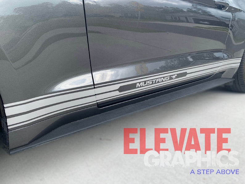 K&ouml;nnte beinhalten: Ein grauer Ford Mustang mit einem schwarzen und grauen Streifenaufkleber an der Seite. Der Aufkleber enth&auml;lt das Wort "Mustang" und ein stilisiertes Pferdelogo. Der Aufkleber stammt von Elevate Graphics, einem Unternehmen, das sich auf Autografiken spezialisiert hat.
