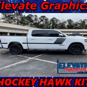Pode incluir: Caminhonete Ram branca com adesivos gráficos de hockey hawk pretos e cinzentos no lado. A caminhonete está estacionada num parque de estacionamento. Os adesivos apresentam as palavras "Hockey Hawk Kit" e o logotipo de um hockey hawk.