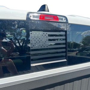 May include: A white pickup truck with a black American flag decal on the rear window. The flag is made of stars and stripes.