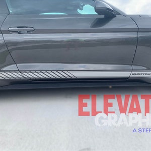 K&ouml;nnte beinhalten: Ein grauer Ford Mustang mit einem grauen und schwarzen Streifenaufkleber an der Seite. Der Aufkleber hat ein sich wiederholendes Muster aus diagonalen Linien. Das Auto hat das Wort "Mustang" an der Seite. Das Auto steht auf einer grauen Oberfl&auml;che.
