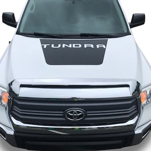 Peut inclure: Un pick-up Toyota Tundra blanc avec un graphisme noir sur le capot qui dit "Tundra" en lettres blanches. Le camion a une calandre et des phares chromés.