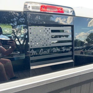 May include: A black truck window with a black and white American flag decal and the words "In God We Trust".