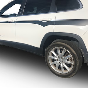 Puede incluir: Jeep Cherokee blanco con rayas laterales negras y una rueda plateada. El coche está aparcado en una superficie blanca.