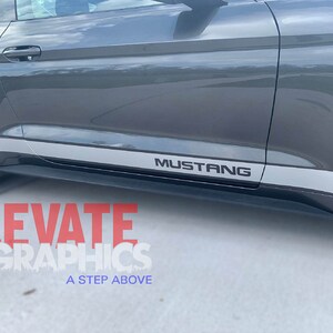 K&ouml;nnte beinhalten: Ein grauer Ford Mustang mit einem silbernen Streifen an der Seite und dem Wort "Mustang" in schwarzen Buchstaben auf dem Streifen.