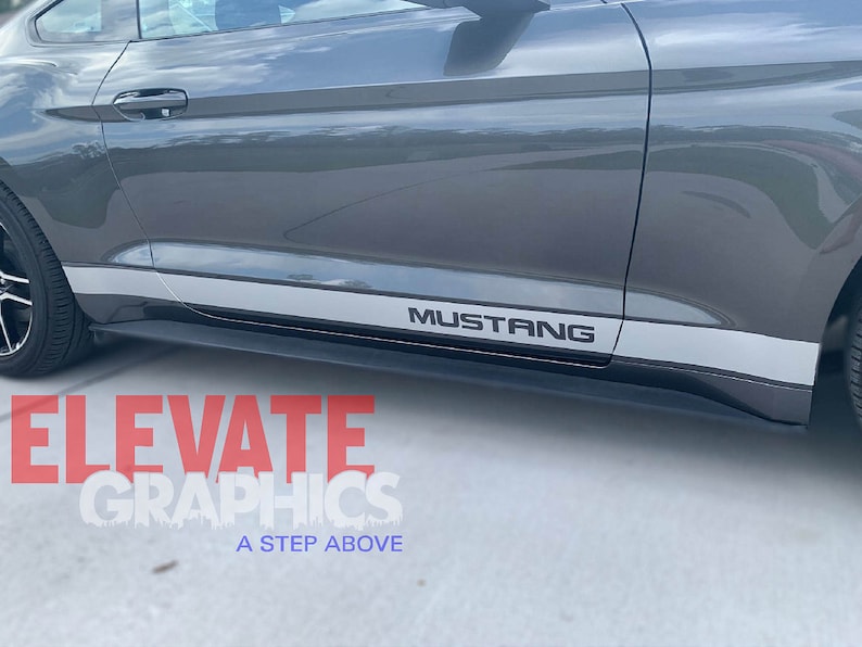 K&ouml;nnte beinhalten: Ein grauer Ford Mustang mit einem silbernen Streifen an der Seite und dem Wort "Mustang" in schwarzen Buchstaben auf dem Streifen.