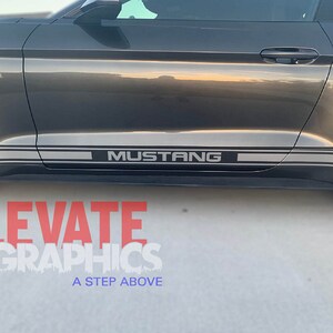K&ouml;nnte beinhalten: Ein grauer Ford Mustang mit einem silbernen Streifen an der Seite des Autos. Der Streifen tr&auml;gt den Schriftzug "MUSTANG" in schwarzen Buchstaben.