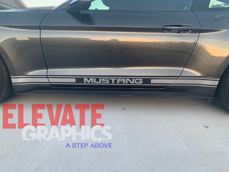 K&ouml;nnte beinhalten: Ein grauer Ford Mustang mit einem silbernen Streifen an der Seite des Autos. Der Streifen tr&auml;gt den Schriftzug "MUSTANG" in schwarzen Buchstaben.