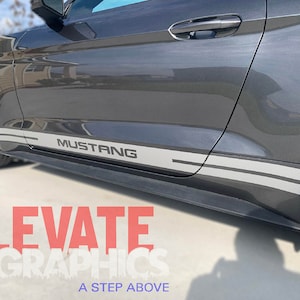 K&ouml;nnte beinhalten: Ein grauer Ford Mustang mit zwei grauen und schwarzen Seitenstreifen und dem Wort "Mustang" in grauer Schrift auf der Seite des Autos.