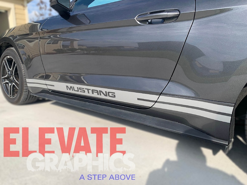 K&ouml;nnte beinhalten: Ein grauer Ford Mustang mit zwei grauen und schwarzen Seitenstreifen und dem Wort "Mustang" in grauer Schrift auf der Seite des Autos.