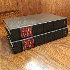 May include: Two hardcover books titled "Institutes of the Christian Religion" by Calvin. The books are dark brown with red labels and gold lettering. The spines read "Calvin". Volume I is stacked on top of Volume II.