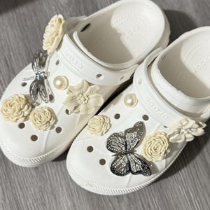 May include: White clogs decorated with cream-colored flowers, pearl accents, and silver butterfly and dragonfly charms. The clogs are a casual style of footwear.