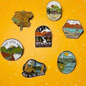 May include: A collection of enamel pins with outdoor and camping themes. Designs include a sign that says "Take a Hike", a jeep with the text "Keep it Simple", and a mountain scene with the text "Life is Better in the Mountains".