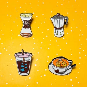 May include: Four enamel pins featuring coffee-related designs. Includes a pour-over coffee maker, a stovetop espresso maker, an iced coffee in a clear cup, and a latte with a heart design. All pins have black outlines and are set against a yellow background.