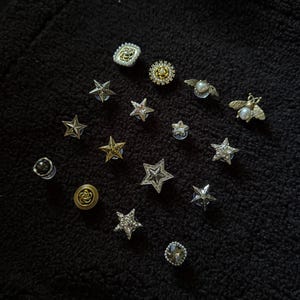 Op de afbeelding: Een verzameling decoratieve spelden en knopen in verschillende vormen, waaronder sterren, bijen en bloemmotieven. De spelden hebben zilveren, gouden en parelaccenten, sommige zijn versierd met kristallen. De items zijn gerangschikt op een zwarte, getextureerde ondergrond.