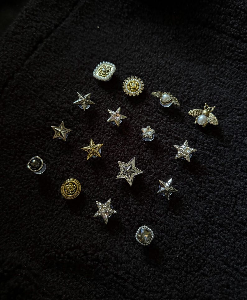 Op de afbeelding: Een verzameling decoratieve spelden en knopen in verschillende vormen, waaronder sterren, bijen en bloemmotieven. De spelden hebben zilveren, gouden en parelaccenten, sommige zijn versierd met kristallen. De items zijn gerangschikt op een zwarte, getextureerde ondergrond.