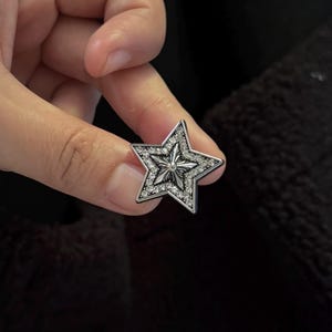 Op de afbeelding: Een zilveren stervormig accessoire met een centraal ontwerp en bezet met kleine, heldere edelstenen. De ster heeft een vijfhoekige vorm en wordt tegen een donkere achtergrond gehouden.